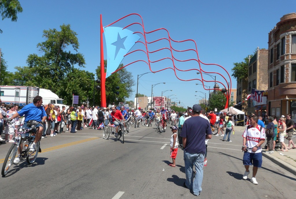 A parade passes under a statue of a Puerto Rican flag in the Paseo Boricua neighborhood of Chicago. The city gifted a matching pair of flag statues to the Puerto Rican neighborhood in 1995. Flickr photo by Emily. /photos/ebarney/2578770769